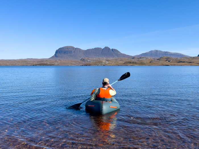 Man paddles packraft on calm waters with the mountain of Suilven in the distance