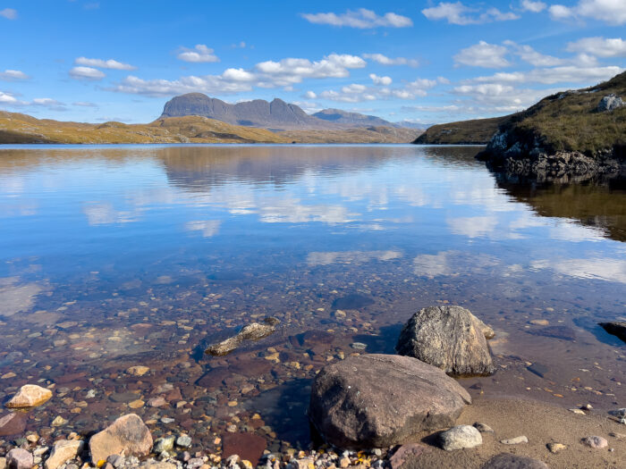 A calm loch with the sky and surrounding mountains reflected