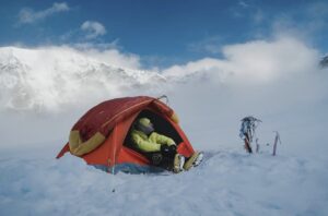 A climber looks out while sitting at the gate of his tent, wrapped in a cloud that slowy recedes to reveal a stunning mountain landscape.