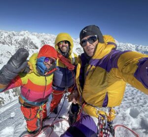 Three climbers stand on a snowy summit in a cold-looking but sunny day.