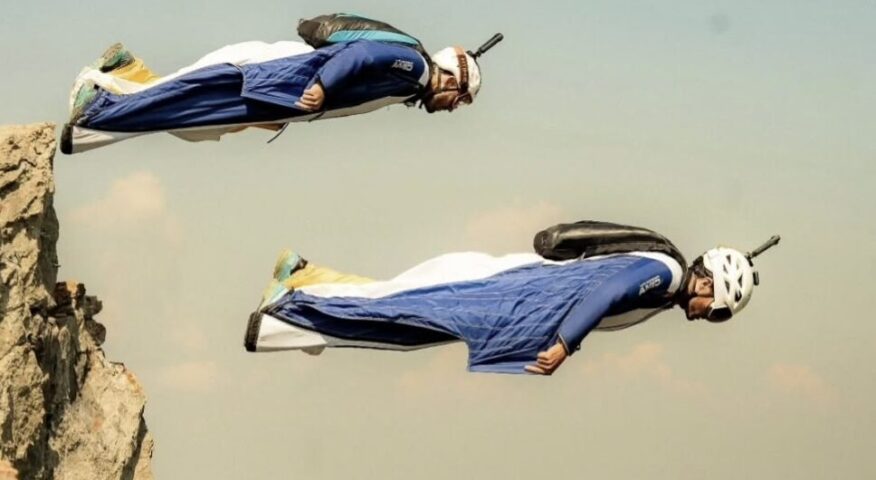 Two base jumpers in matching wingsuits take off from a rocky outcrop.