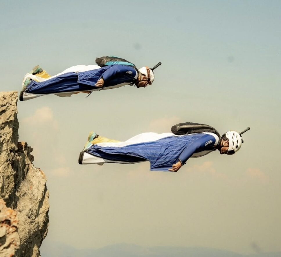 Two base jumpers in matching wingsuits take off from a rocky outcrop.