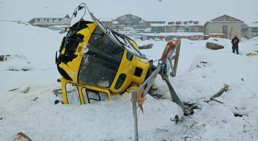 A crashed helicoper on snowy ground with buildings of a village behind.