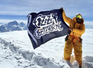 A climber on the flat top of Cho Oyu, waves a flag of Ozzy osbourne, Everest in background.