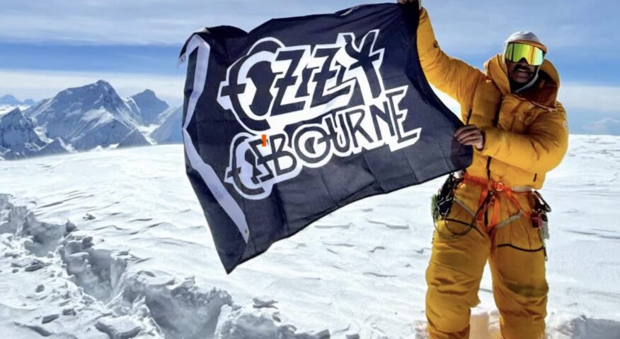 A climber on the flat top of Cho Oyu, waves a flag of Ozzy osbourne, Everest in background.