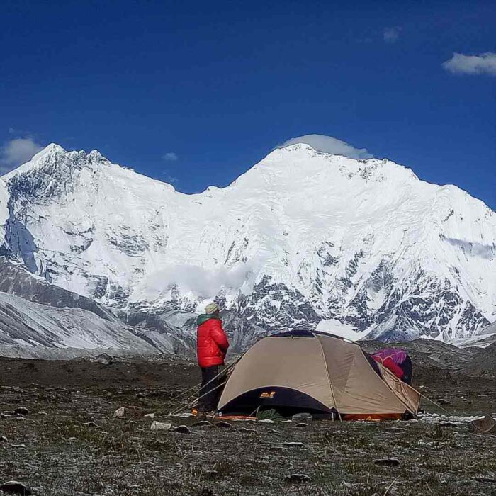 A trekker by a tent on a green meadow, Everest and Lhotse in background.