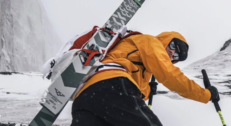 Jim Morrison with skis on his back climbing up a steep snow slope.