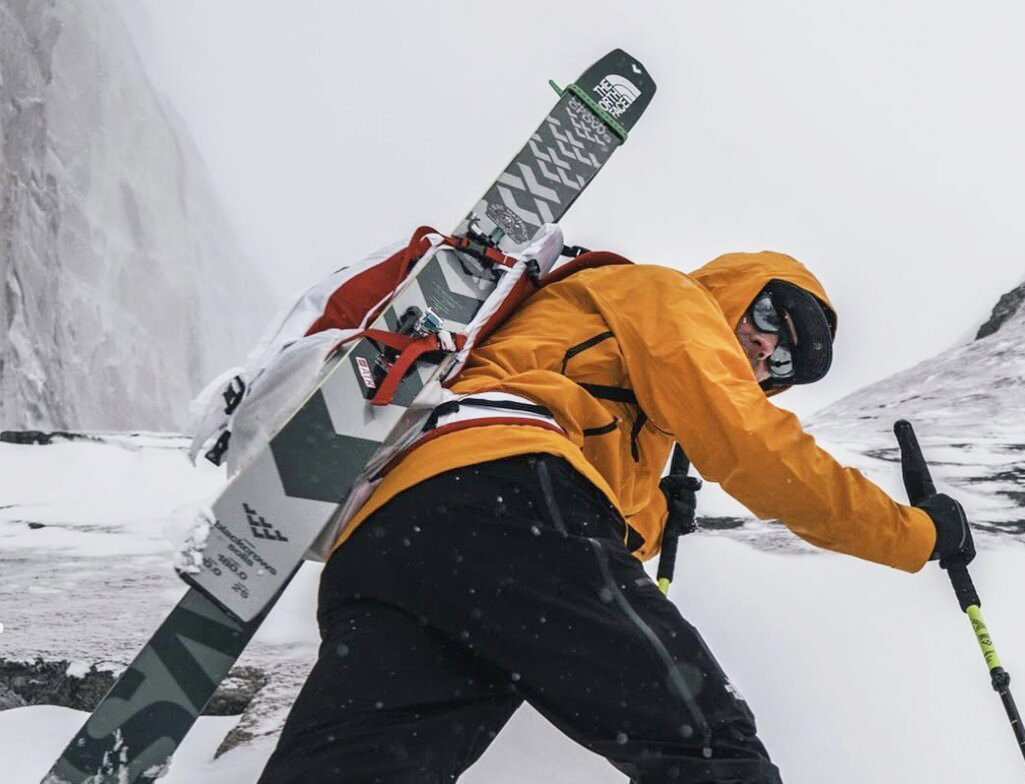 Jim Morrison with skis on his back climbing up a steep snow slope.