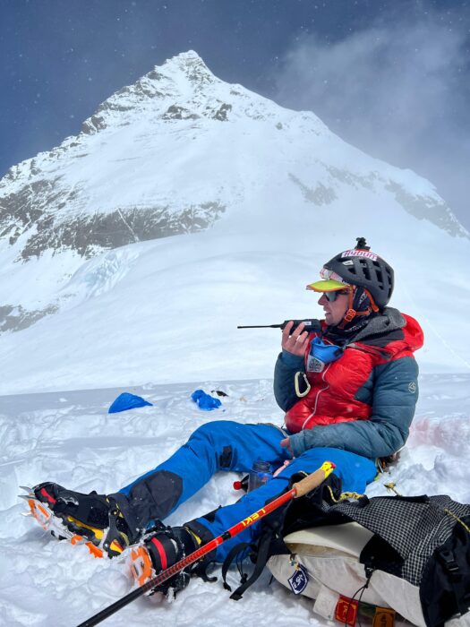 Tyler Andrews speaks on the radio, sitting on the snow at the South Col, with the summit section of Everest behind him. 