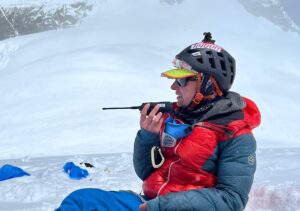 Tyler Andrews sits on the snow and talks on the radio, with the summit pyramid of Everest behind him,