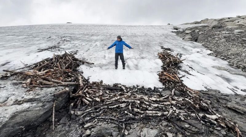 A researcher stands on top of the melting ice that has revealed the wooden structures. These can be seen coming out of the ice