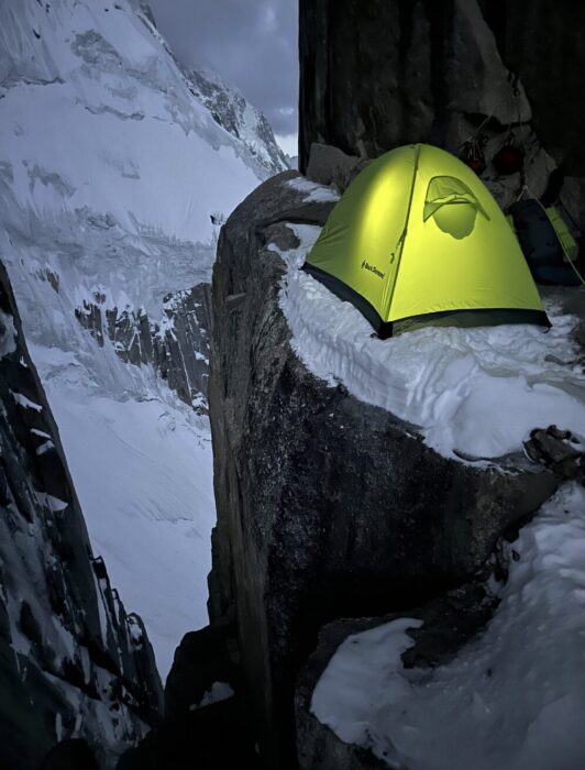 A tent lit from the inside, perched on a ledge in a snow and rock face.