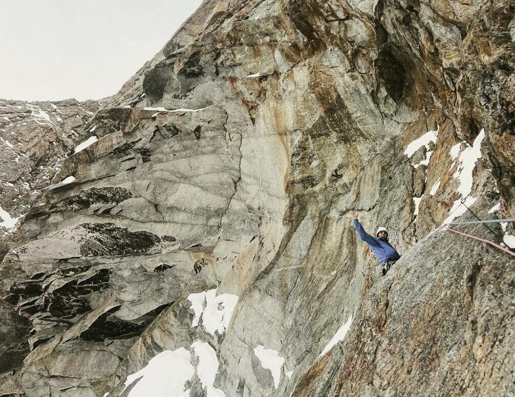 A climber cheers from a vertical granite face, with some patches of snow.