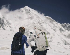 two climbers look at a mountain, one of them pointing to it with his walking pole.