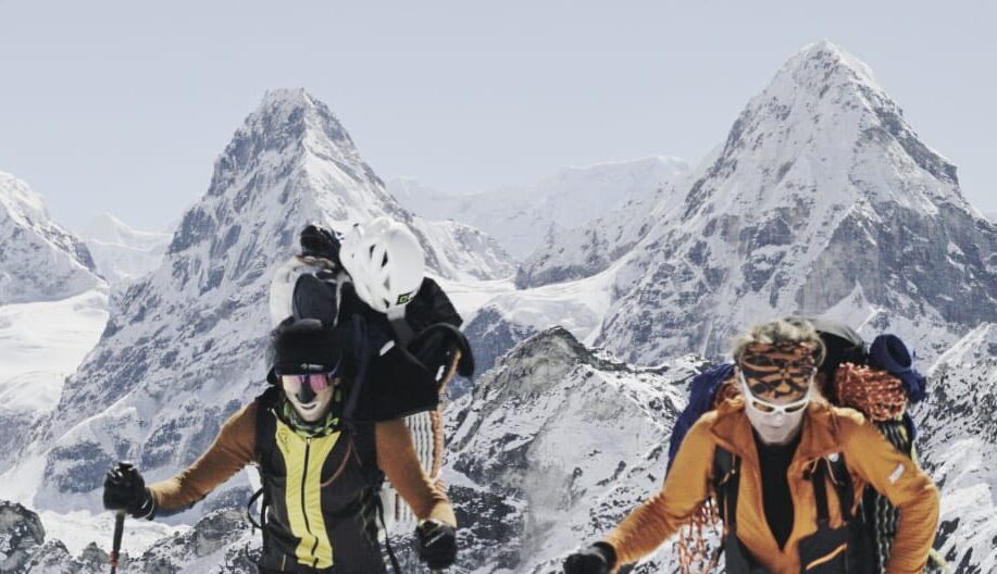 Climbers with heavy backpacks and walking poles, with spiky peaks behind them.
