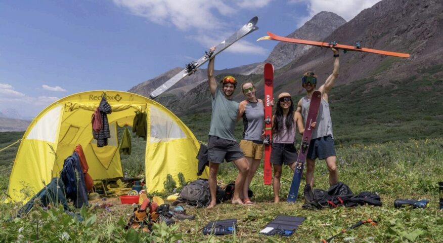 Four skiers in shorts and t-shirts by a tent on a grassy meadow.