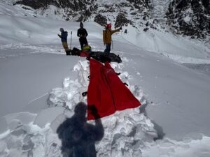 Four rescuers on a snowy surface where they have dug avalanche victims and covered them with a red blanket.
