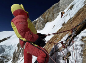 A climber belays another on a big mixed face, with high-altitude suits.