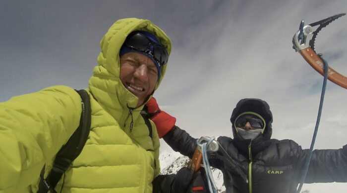 The climbers rise their arms in triumph on a summit, with a cloud behind them. 