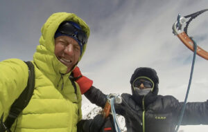 two climbers raise their arms in triumph from a summit