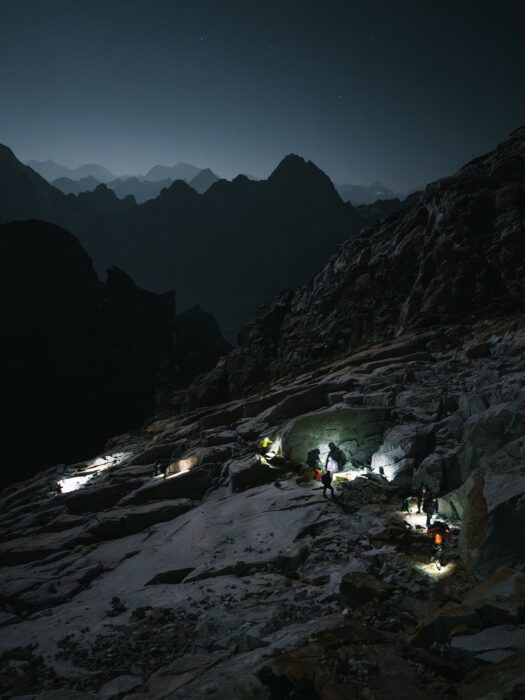 Climbers progressing on scree and snow before dawn with headlamps on.