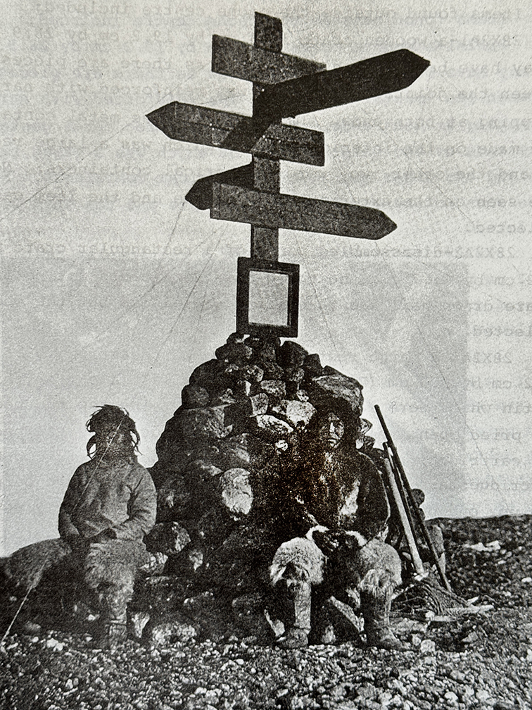old black and white photo of a signpost with two Inuit in front of it
