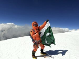 A climber in high altidue gear on a flat snow summit, mountains wrapped in clouds in the background.