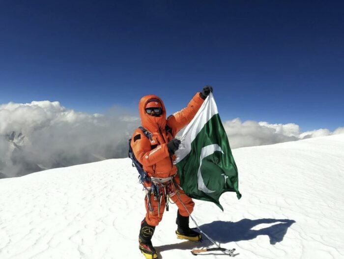 A climber in high altidue gear on a flat snow summit, mountains wrapped in clouds in the background.