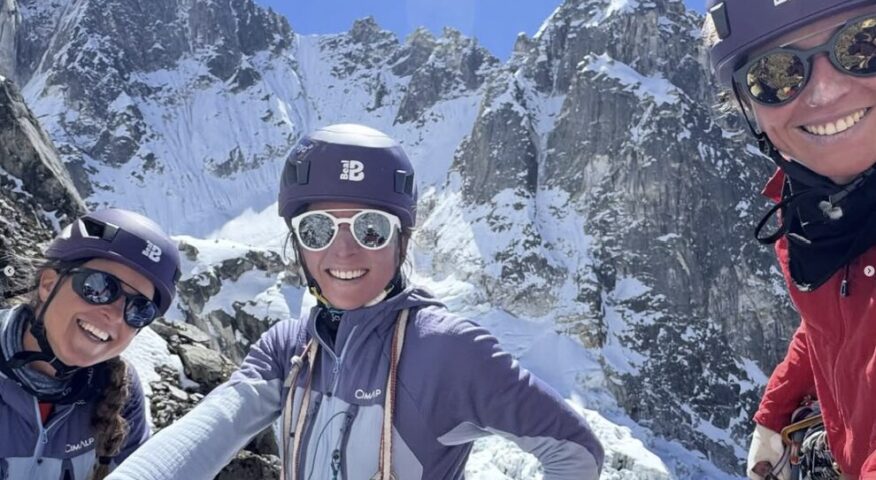 Three female climbers with helmets and sunglases, with rocky spire rising from the snow behind.
