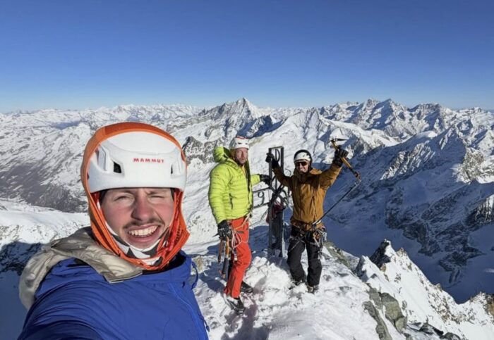 Climbers smile on a snowy summit in a clar, cold day. 