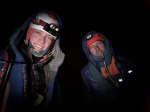 two climbers with warm clothes, helmets and headlamps smile in the night.