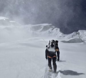 Climbers up a snow slope clipped to afixed rope with wind rising clouds of snow.
