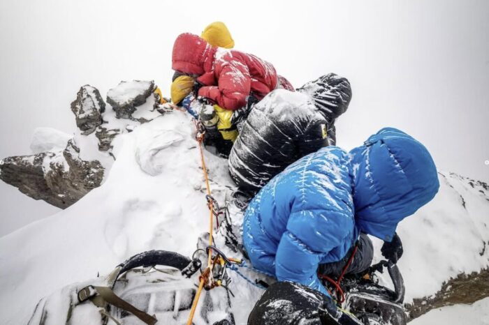Climbers sitting on iced surface, looking down as snow covers them. 