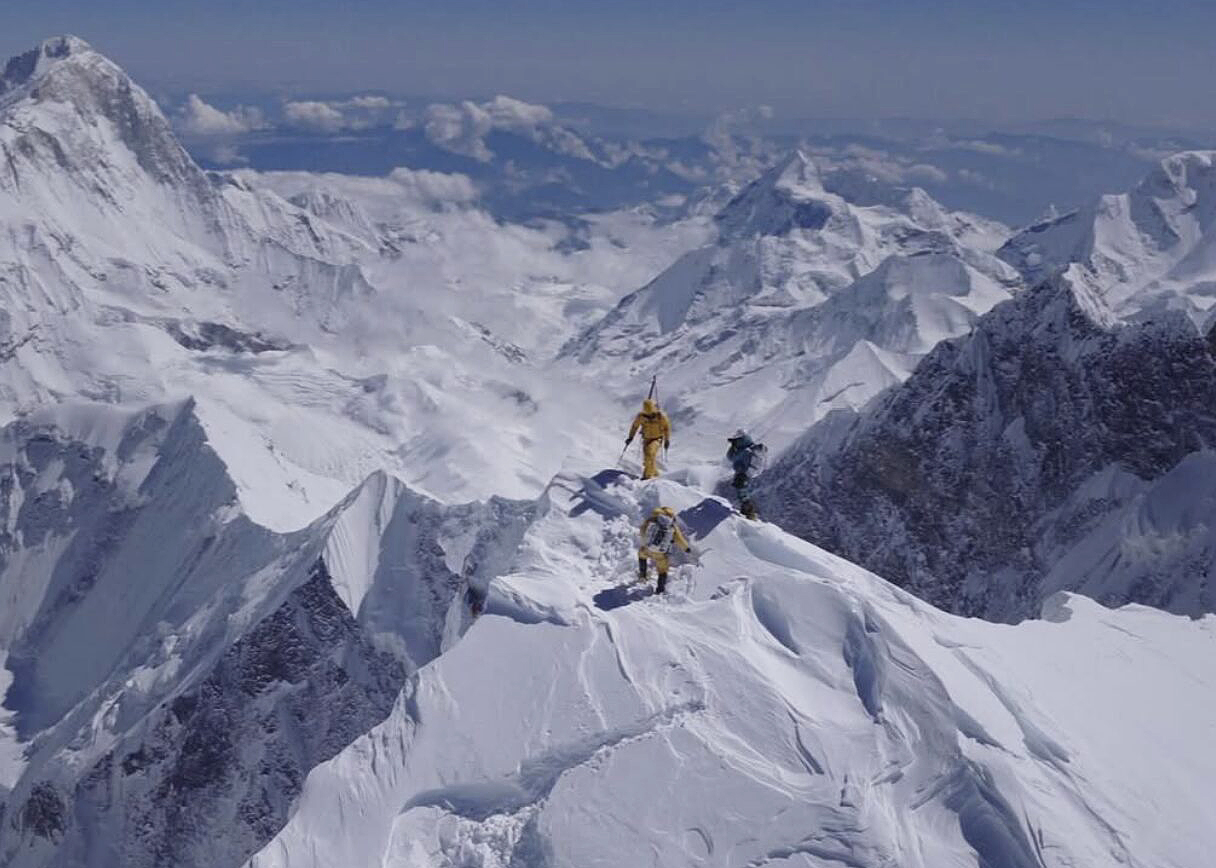 A small group of climbers on the summit of Everest, surrounded by mountains.