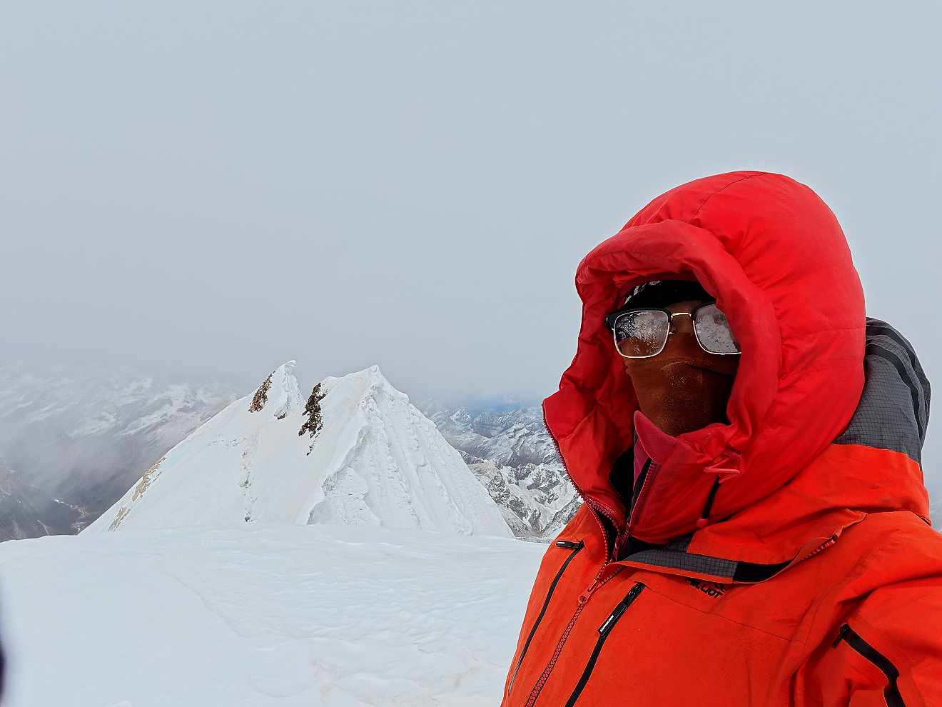 A Chinese climber on a snowy summit, with a slightly lesser point behind him, in a cloudy day.