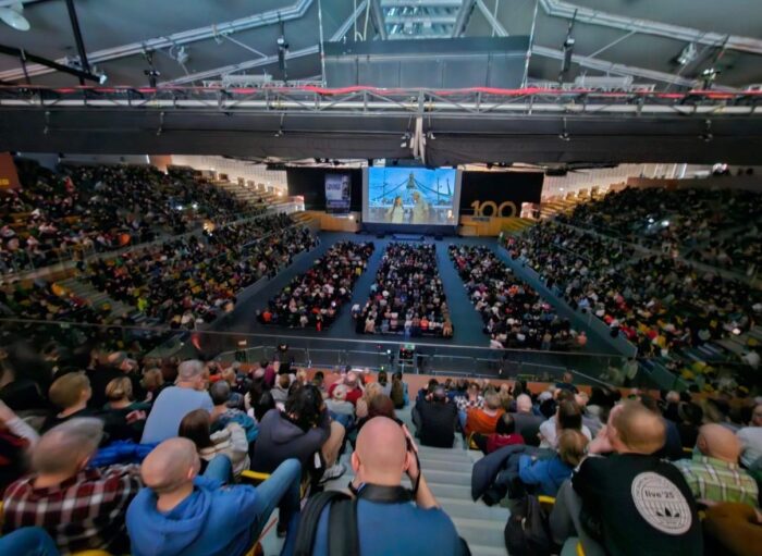 A Basquetball stadioum plenty of people seeing a lecture on a screen.