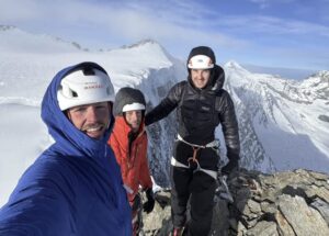 Three climbers smile on an alpine summit.