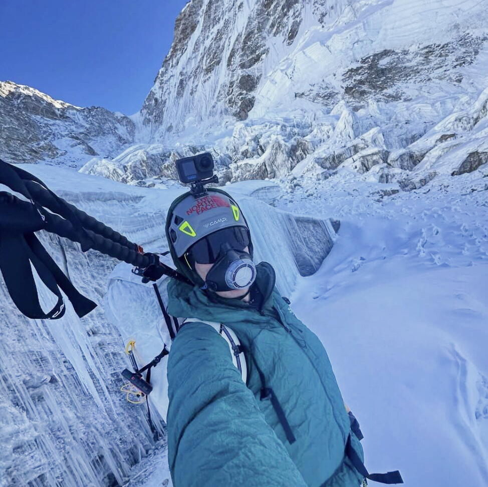 A climber with an air-warming mask takes a selfie in front of a technical mountain face.