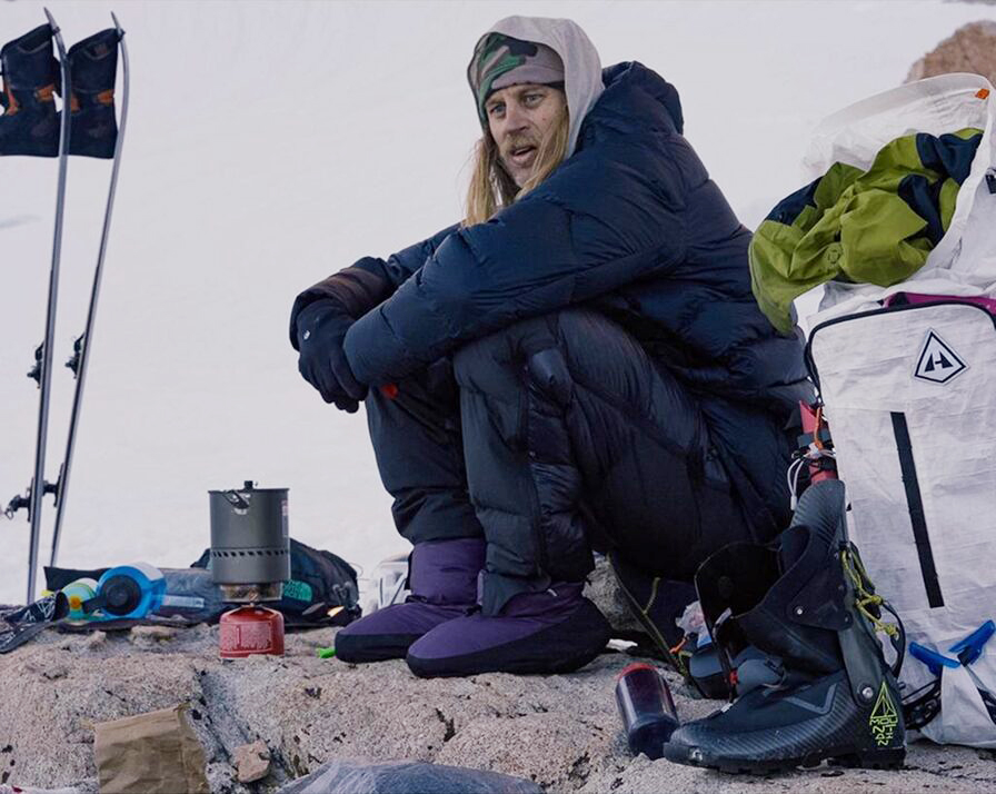 A climber looking cold and tired at a bivouaq, with a little stove in front.