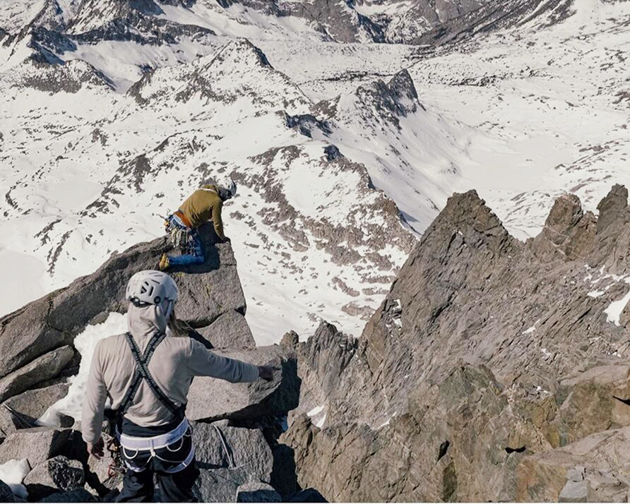 Two climbers on a rocky ridge with snowy terrain below.