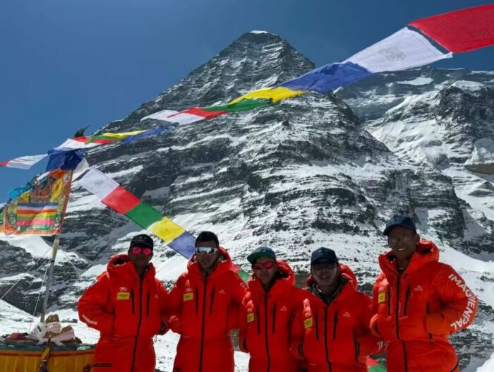 Sherpa climbers in red suits with the Dhaulagiri's Eiger and prayer flags behind them. 