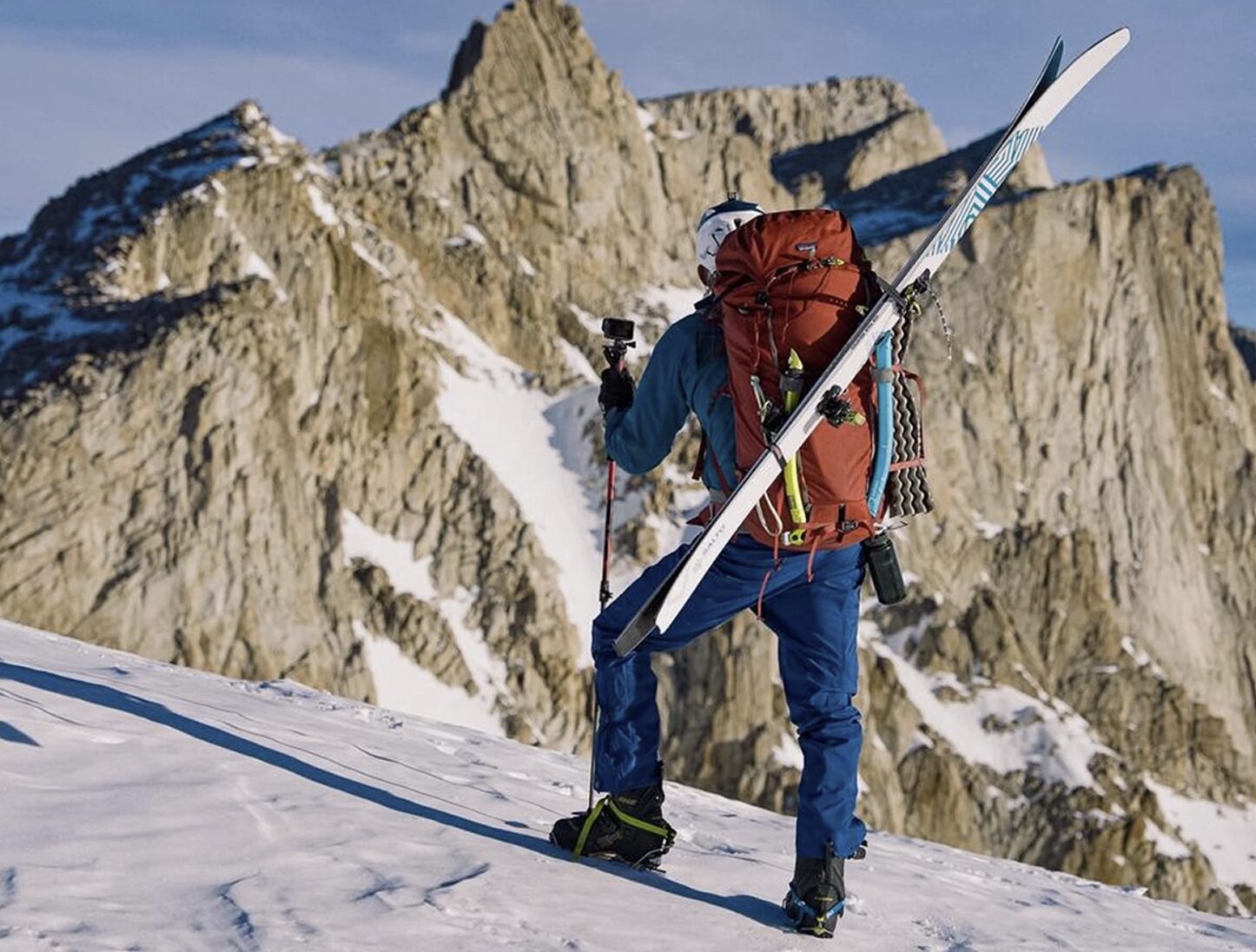 A climber stands on a snowy summit with a rocky peak in front of him, carrying skis and ice-axes on his backpack.