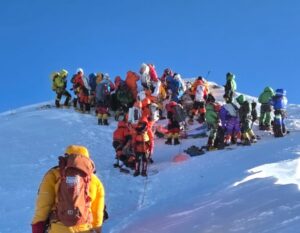About 20 climbers stand up on a crowded Everest summit in a sunny day.