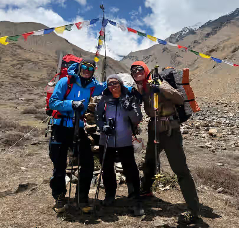 Three climbers smile and pose by a chorten in a barren landscape.