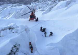 A helicopter about to land on top of a serac, a group of climbers , one of them hanging from a rope, below.