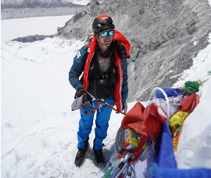 Andrews stands on glacier terrain with his packpack on the snow by some prayer flags.