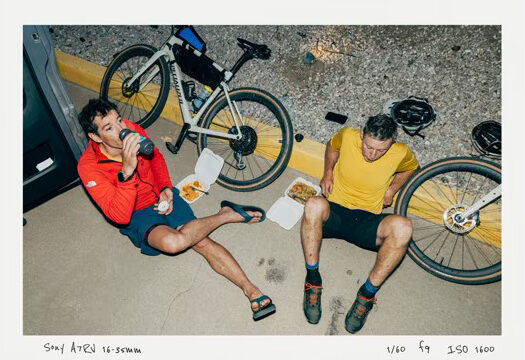 Two men with bikes sitting on the road eating takeout