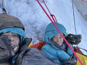 Two female climbers with jackets and hoods on, at a portaledge on an ice wall.