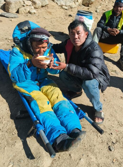 A worker gives a bowl of soup to an injured Sherpa on a stretcher. 
