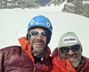 Two climbers with helmets on smile at the camera, a snow slope leading to a col behind them.